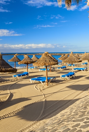 A view over a beach in Los Cristianos in Tenerife with golden sand, blue skies and grass parasols.